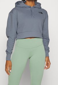 Felpa grigia accorciata con cappuccio e logo, abbinata a leggings a vita alta di un verde chiaro. Materiale morbido con texture a costine sui polsini.
