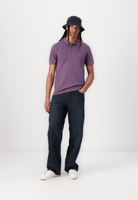 Purple polo shirt with short sleeves, a small logo, paired with dark wide-leg jeans and white sneakers. The model wears a black bucket hat.