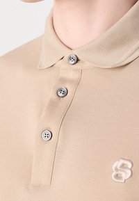 Polo beige à trois boutons avec un petit logo brodé blanc en forme de "S" sur la poitrine, porté par une personne montrant son cou et son menton.