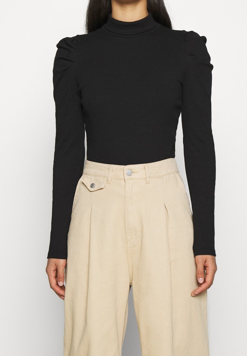 Body a maniche lunghe nero con collo a dolcevita e spalle imbottite, abbinato a pantaloni beige a vita alta plissettati con tasche laterali.