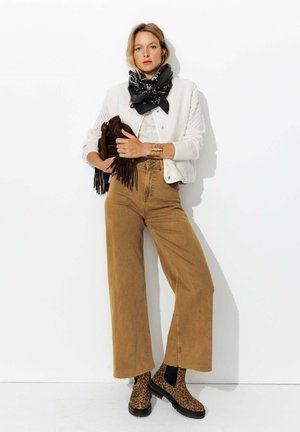 Pantalons larges kaki associés à une veste blanche texturée, une écharpe noire et des bottines à imprimé léopard. Tenant une pochette frangée marron.