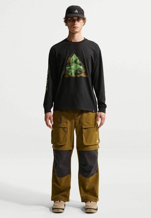 Νέος άνδρας που φοράει μαύρο πουλόβερ με γραφικό πράσινο τρίγωνο, δίχρωμο καφέ cargo παντελόνι, μπεζ αθλητικά παπούτσια και μαύρο καπέλο.