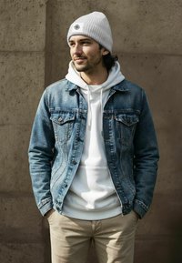Denim jacket with front pockets, light blue color, paired with a white hoodie and beige pants. A gray beanie tops the look, featuring a logo.