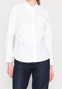 Femme portant une chemise blanche cintrée à manches longues avec une poche poitrine et un jean bleu foncé, debout devant un fond uni.