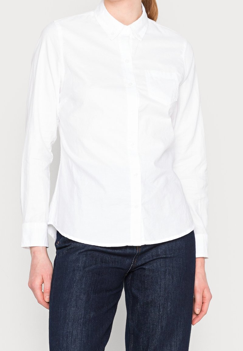 Femme portant une chemise blanche cintrée à manches longues avec une poche poitrine et un jean bleu foncé, debout devant un fond uni.