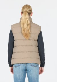 Beige pufferväst med horisontell quiltad design, hög krage och en slät yta, kombinerad med en mörk långärmad tröja och ljusblå jeans.
