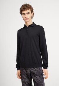 Polo preto de manga longa com um fecho de três botões, gola clássica, punhos com ribetes e uma textura de malha suave. Combinado com calças estampadas.