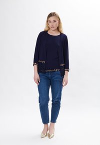 Cardigan blu con dettagli a occhiello e accenti circolari ai polsini, abbinato a una maglietta blu e jeans. Tacchi a punta dorati.