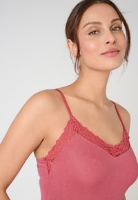 Camisole rose avec des détails en dentelle le long de l'encolure et des bretelles, présentant une texture lisse et une coupe ajustée. Bretelles spaghetti réglables.