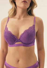 Fioletowy koronkowy biustonosz z głębokim dekoltem, skomplikowanymi wzorami kwiatowymi, regulowanymi ramiączkami oraz gładkim pasem pod biustem z wytłoczonym logo.