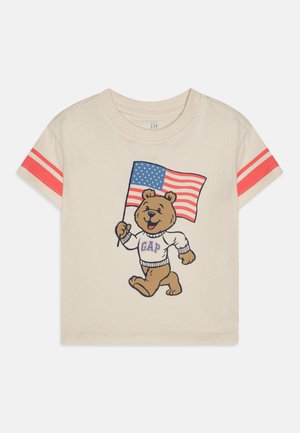Tejszínből készült, rövid ujjú póló, amelyen egy rajzfilmszerű medve látható, aki egy amerikai zászlót tart. A póló piros csíkos ujjaival rendelkezik; a grafikán a medve pulóverén a "GAP" felirat szerepel.