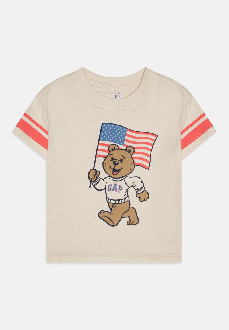 Krémové tričko s krátkymi rukávmi, na ktorom je obrázok karikatúrneho medveďa, ktorý drží americkú vlajku. Rukávy sú pruhované červenou farbou; grafika zobrazuje nápis "GAP" na medveďovom svetri.