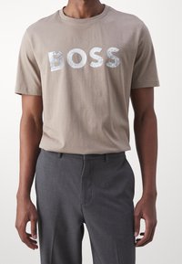 Muž v béžovém tričku s metalickým nápisem "BOSS" a šedých společenských kalhotách, stojící na bílém pozadí.