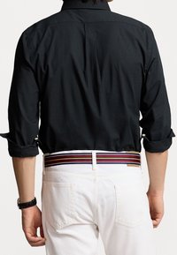 Svart skjorta med upplagda ärmar, i kombination med vita byxor som har ett flerfärgat randigt bälte. Bakifrån, som visar skjortans ryggveck och manschett.