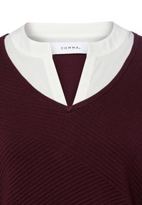 Sweter w kolorze bordowym z dekoltem w serek, o fakturowanej powierzchni, nałożony na białą koszulę z kołnierzem typu notch oraz widoczną etykietą COMMA przy dekolcie.