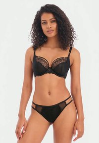 Donna con capelli ricci che indossa un set di lingerie in pizzo nero, in piedi contro uno sfondo bianco uniforme, mentre guarda verso la fotocamera.