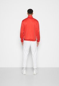 Chaqueta deportiva roja con cuello alto, combinada con pantalones deportivos blancos. Tejido suave, detalles en blanco que contrastan en las mangas, diseño minimalista.