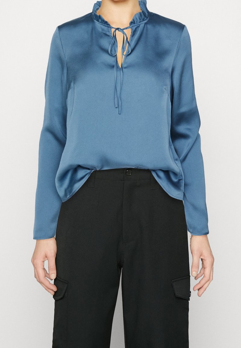 Blusa di raso blu con maniche lunghe, scollatura a ruches, dettaglio con fiocco davanti, abbinata a pantaloni cargo neri con tasche laterali.