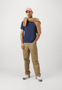 Marine t-shirt med et hvidt logo, beige bukser og en brun sweater draperet over skuldrene. Lyserød kasket og hvide sneakers med grå detaljer.