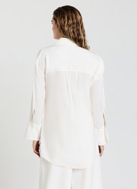 Blusa bianca a maniche lunghe in tessuto leggero, con piega sul retro, orlo arrotondato e polsini con bottoni dotati di aperture a fessura.