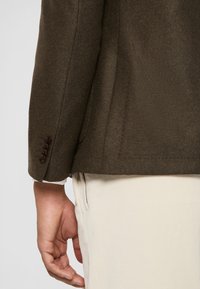 Sacou din lână maro, cu țesătură texturată, croială ajustată și detalii cu cinci nasturi la manșete. Pantaloni bej deschis vizibili sub sacou.