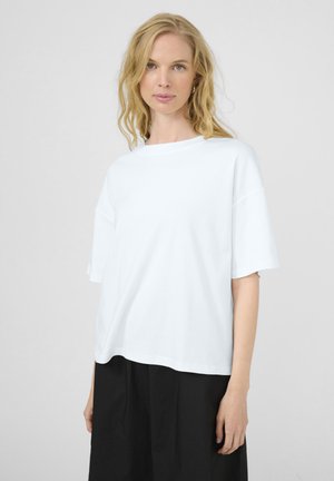 Kobieta o blond włosach, ubrana w luźną białą koszulkę z krótkim rękawem i czarne spodnie, stojąca na tle jednolitego jasnego tła.