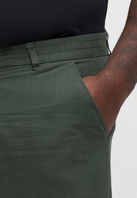 Pantalon vert en chevrons avec une poche latérale, doté d'une ceinture robuste et d'une coupe décontractée. Le matériau semble doux et durable.