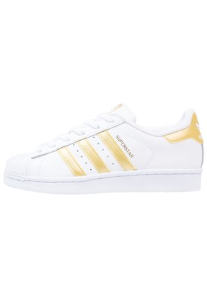Basket blanche Adidas Superstar avec des bandes dorées, un onglet de talon doré et le texte "Superstar" sur le côté, présentée en profil latéral face à droite.