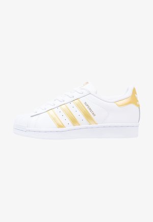 Hvid Adidas Superstar sneaker med guldstriber, gyldent hælstykke og tekst "Superstar" på siden, vist i sideprofil mod højre.