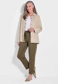 Beige suède shirtjack, witte top, olijfgroene broek met riem en lichte sandalen. Voorzien van knoopsluitingen en zijsplitten op de broek.