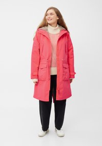 Parka impermeable rosa con capucha, dos bolsillos frontales, cordones ajustables y un forro interno suave, combinada con un suéter crema y pantalones negros.