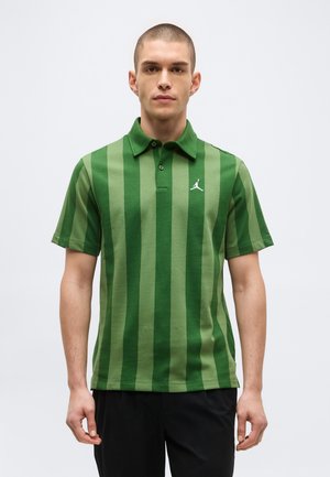 Férfi zöld, függőleges csíkos, rövid ujjú galléros pólót és fekete nadrágot visel, sima világos háttér előtt áll.