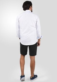 Weißes Langarmhemd mit umgeschlagenen Manschetten, kombiniert mit schwarzen Shorts. Das Modell trägt blaue Schuhe. Glatte Textur, abgerundeter Saum und Knopfkragen-Detail.