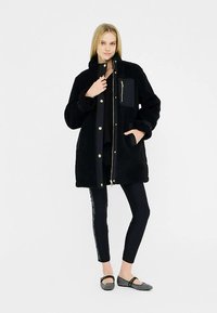 Sort, fluffy overtøj med lynlås og trykknaplukning, stor frontlomme og guldfarvede detaljer. Båret over sort top og bukser.