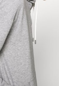 Grijze hoodie van zacht, gestructureerd materiaal met een kangoeroezak, witte trekkoorden en metalen uiteinden. Strak, casual ontwerp.