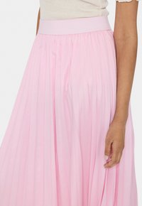 Jupe plissée rose pâle avec une large taille élastique. Le tissu a un léger éclat, créant une texture douce et fluide.
