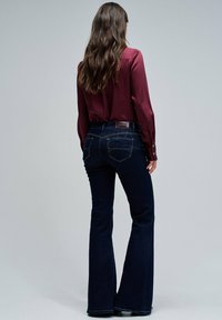 Mujer con largo cabello castaño ondulado lleva una blusa burdeos y jeans oscuros de campana, de pie contra un fondo gris claro liso.