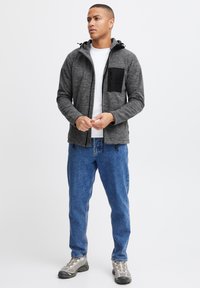 Grå zip-up jakke med en sort brystlomme, parret med blå jeans. Jakken har en hætte, justerbare snore og tekstureret stof.