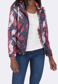 Floral bomber jacket in dark blue with red and white leaf patterns. Features a zip front, hood, and elastic cuffs. Worn over a white top.