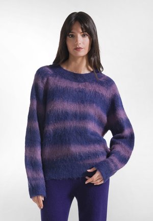 Kvinde med mørkt hår iført en blød lilla og pink stribet sweater og matchende lilla bukser, står foran en ensfarvet lys baggrund.