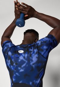 Man in een blauw en zwart sportshirt tilt een blauwe dumbbell van 1,5 kg boven zijn hoofd met beide handen tegen een effen grijze achtergrond.