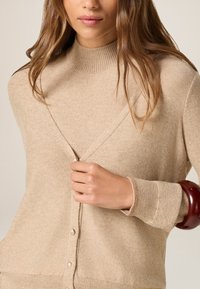 Cardigan in maglia beige con colletto alto, caratterizzato da un design a V, bottoni perlacei e polsini a costine. Texture liscia con un sottile luccichio.
