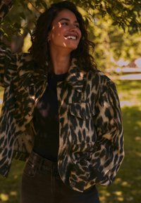 Tekoturkistakki musta- ja ruskeapilkullisella leopardi kuosilla, jossa on kauluskrae, nappisuljenta ja kaksi etutaskua.