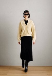 Un cardigan en tricot jaune clair avec des boutons, porté sur une robe midi noire à côtes et associé à des bottines noires. Le modèle se tient contre un fond neutre.