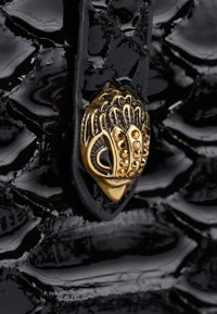 Zwart textuurmateriaal met een glanzende afwerking, voorzien van een decoratieve sluiting in goudkleur met verfijnde details en kleine edelstenen.