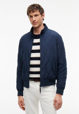 Uomo che indossa una giacca trapuntata blu navy sopra un maglione a righe bianche e nere con pantaloni bianchi, in piedi davanti a uno sfondo bianco uniforme.