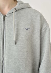 Grå dragkedja-hoodie i mjukt material, med en tvåfärgad dragsko och en liten mörk logotyp på bröstet. Enkel, avslappnad design.