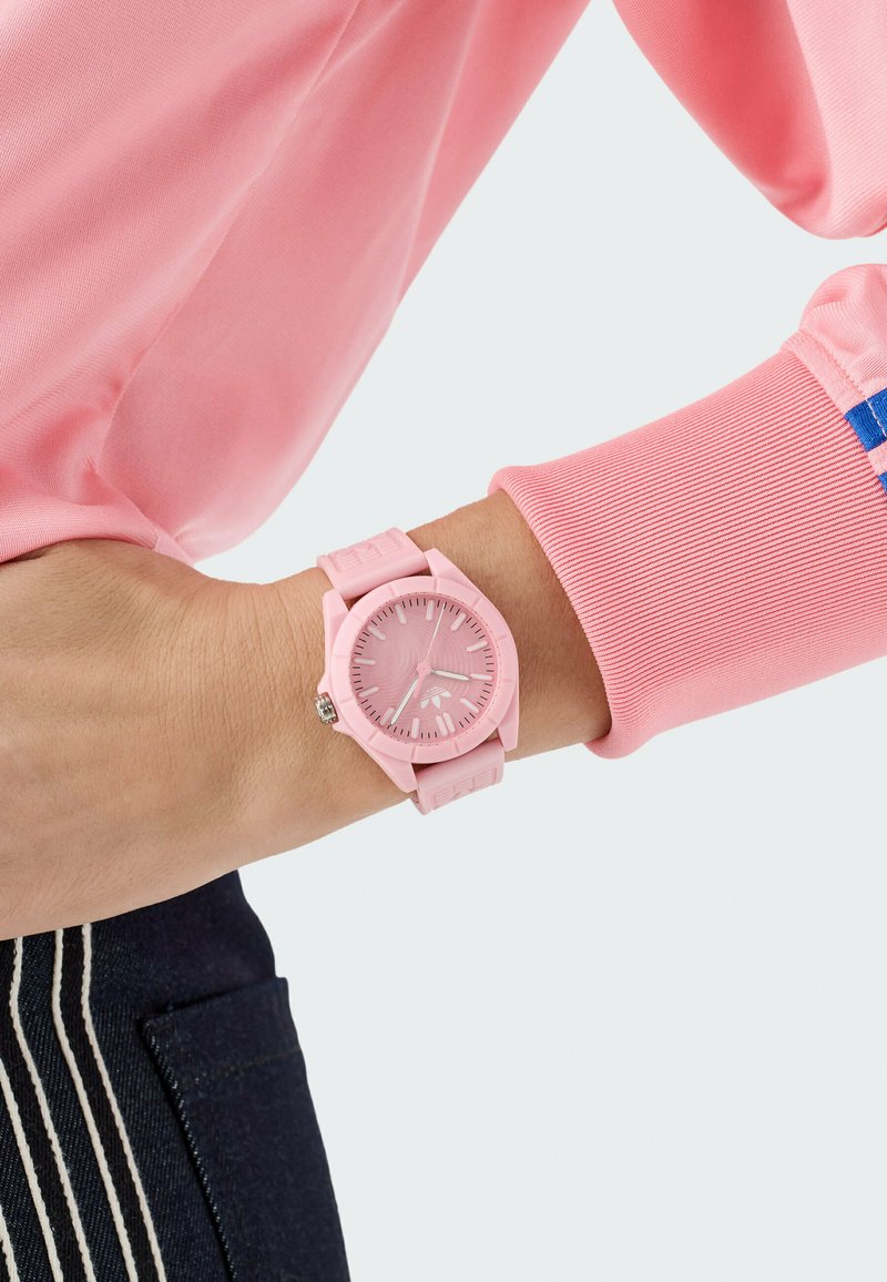 Montre-bracelet en silicone rose avec un cadran rond, comportant un cadran texturé et des marqueurs d'heure simples. Portée à un poignet avec une manche rose.