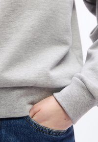 Main dans la poche avant d'un jean bleu, personne portant un sweat-shirt gris clair avec une manche retroussée.