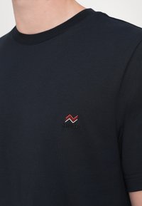 T-shirt à manches courtes de couleur marine en tissu doux, avec un petit logo zigzag rouge et blanc brodé sur la poitrine gauche.